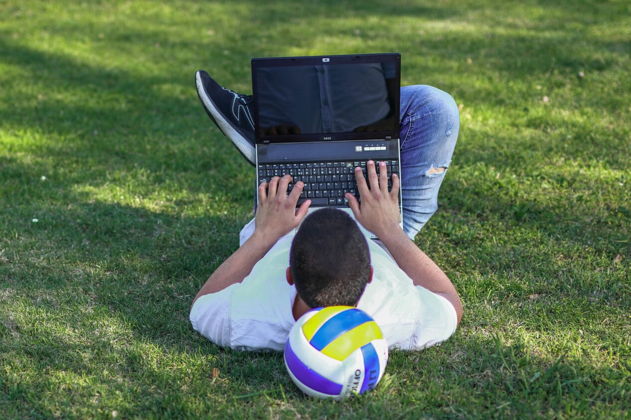 Pojke ligger på gräsmatta med dator i knät och händerna på tangentbordet.